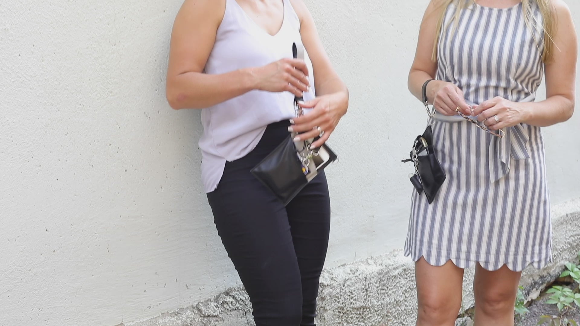 A person with a  black vegan leather phone pouch with a black vegan leather magnetic wristlet key chain and a black removable three pocket wallet and black lip balm holder set. Another Person  with a black and gray vegan leather bracelet, keychain, three pocket wallet, and a black lip balm holder used as a fanny pack belt bag. 